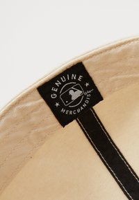 Beige stoffen cap binnenkant met een zwart stoffen label. Het label bevat witte tekst met de woorden "ECHTE MERCHANDISE" en een silhouet.