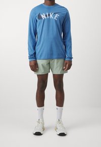 Muž oblečený v modrém dlouhém tričku Nike, světle zelených šortkách Nike, bílých ponožkách Nike a bílých sportovních teniskách stojí na jednobarevném pozadí.