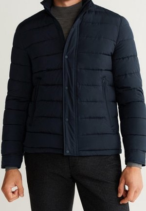 Homme portant une doudoune matelassée bleu marine sur un col roulé gris, assortie à un pantalon à carreaux foncé.