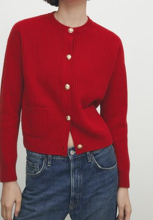 Cardigan tricoté rouge côtelé avec boutons dorés et poches avant, porté avec un jean bleu taille haute, visible du cou aux hanches.