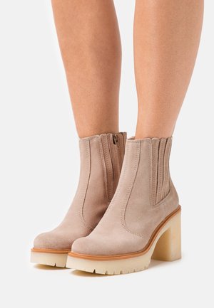 Botines de ante beige con un tacón grueso y translúcido, detalle de ribete en los laterales y cremallera lateral para facilitar su uso. Diseño moderno y texturizado.