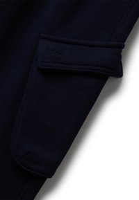 Detailní záběr na tmavě modrou látku s obdélníkovou kapsou, která má klopu a jemně embosované kruhové logo.