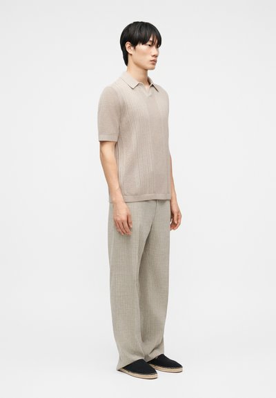 Άνδρας όρθιος, φοράει μπεζ πλεκτό κοντομάνικο πόλο, χαλαρά ανοιχτόγκρι παντελόνια και μαύρα παπούτσια slip-on, κοιτώντας προς τα δεξιά σε απλό φόντο.