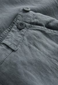 Tissu gris avec une texture douce, deux boutons noirs et un détail de poche cousu. Le matériau semble léger et légèrement froissé.