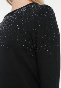 Pull noir en tricot avec des strass argentés dispersés sur les épaules. Le design comprend un col rond et un tissu texturé.