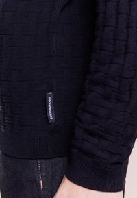 Gros plan sur la manche en tricot texturé foncé et l'ourlet latéral d'un pull, avec une petite étiquette de marque Emporio Armani sur un jean en denim foncé.