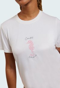 Vit bomull t-shirt med en rosa sjöhäst-illustration och texten "Coastal Wanderer" i närheten. Korta ärmar och rundad halsringning.