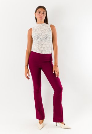 Donna in piedi con top bianco in pizzo senza maniche, pantaloni fucsia aderenti e tacchi alti bianchi a punta su sfondo bianco.