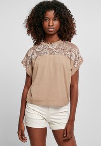 Lys brun t-shirt med blonderoverlay på skuldrene, korte ærmer og en løs pasform. Sammen med hvide, opfoldede shorts.