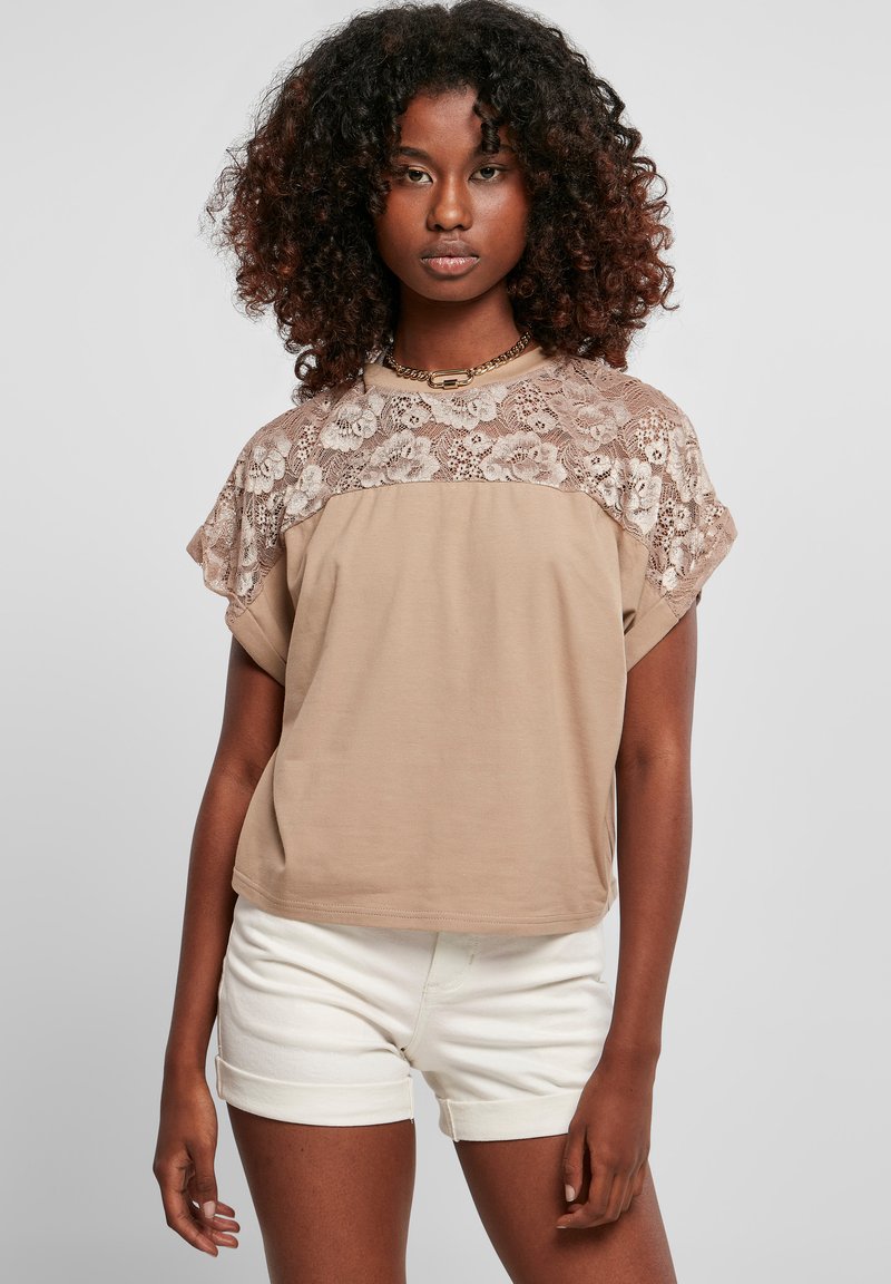 Lys brun t-shirt med blonderoverlay på skuldrene, korte ærmer og en løs pasform. Sammen med hvide, opfoldede shorts.