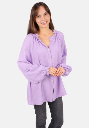 Jeune femme souriante, portant une blouse ample lavande à boutons avec des poignets à volants et un jean gris foncé, se tenant devant un fond blanc.