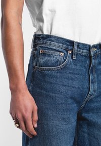 Blue denim jeans with a five-pocket design, copper rivets, and a straight leg. The texture is slightly worn with a medium wash finish.