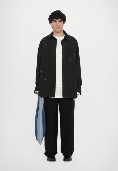 Chemise noire à boutons avec éclaboussures de peinture, T-shirt blanc en dessous, pantalon noir ample et un sac en denim bleu et gris drapé sur la main.