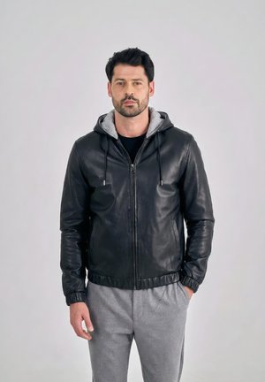 Mand iført sort læder-zip-hoodie over grå joggingbukser, stående mod en ensfarvet lys baggrund, hænder afslappet ved siderne.