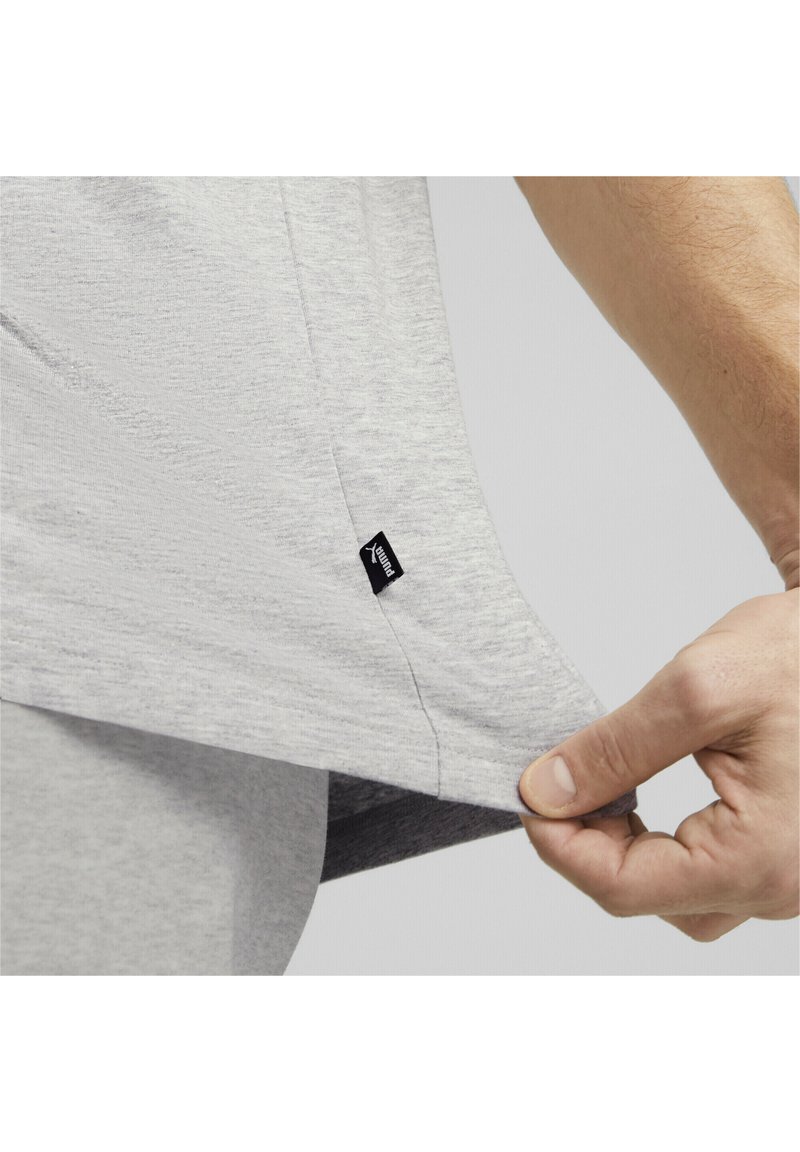 Camiseta deportiva de color gris claro con una tela texturizada. Una mano sostiene el dobladillo inferior, revelando una pequeña etiqueta negra cosida en el lateral.