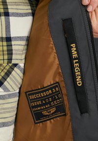Padded jacket featuring a brown interior, gray exterior, and a plaid-patterned fabric. Label includes details in black and gold.