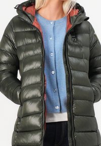 Chaqueta acolchada en verde oscuro con un acabado brillante y un interior naranja contrastante, que presenta un cierre de cremallera y una capucha. Debajo, un cárdigan azul con botones.