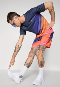 Sportoutfit mit einem navy-blauen zu orangefarbenen Farbverlauf kurzärmligen Shirt und mehrfarbigen Shorts, ergänzt durch weiße Schuhe und Tattoos.