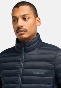 Navyblauwe pufferjack met horizontale stiksels, hoge kraag en volle rits. Bevat het Timberland-logo in lichtblauw op de linkerborst.