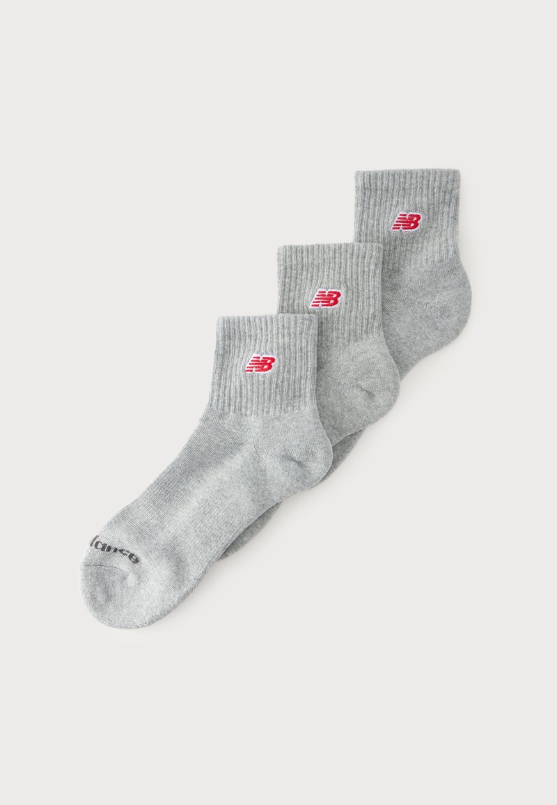 Trois paires de chaussettes grises à la cheville avec des bords-côtes et des logos New Balance rouges sont empilées sur un fond blanc.