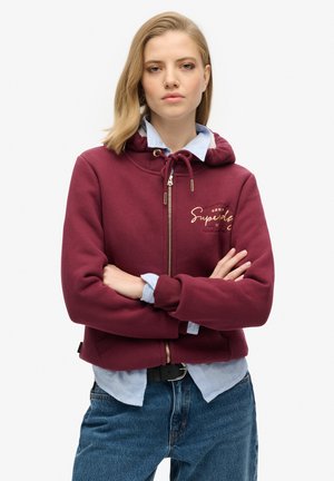 Haut de sweat à capuche zippé bordeaux avec logo doré, capuche à cordons et poignets côtelés, associé à une chemise à col bleu clair et un jean noir.