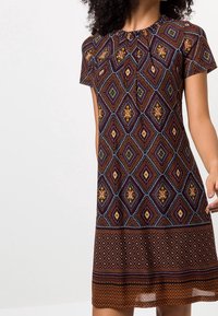 Femme portant une robe à manches courtes avec des motifs géométriques en losange et bordures marron, orange et bleu, se tenant contre un fond blanc uni.