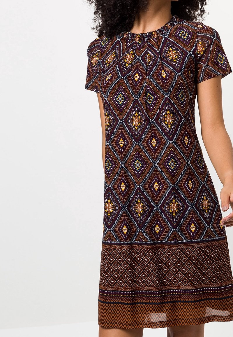 Femme portant une robe à manches courtes avec des motifs géométriques en losange et bordures marron, orange et bleu, se tenant contre un fond blanc uni.