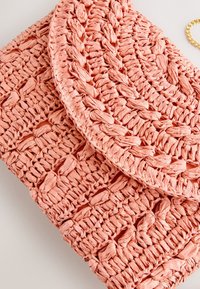 Woven coral pink handbag with a flap closure and chunky braided texture, set against a light background with a gold chain partially visible.
