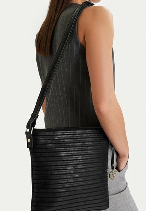 Mujer con top sin mangas de rayas oscuras que lleva un bolso de hombro negro con textura y una correa larga ajustable en la espalda.