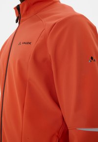 Männersoftshelljacke in Orange mit einem schwarzen Reißverschluss und Logo, ausgestattet mit einem hohen Kragen und glatter Textur. Reflektierende Akzente an den Ärmeln.