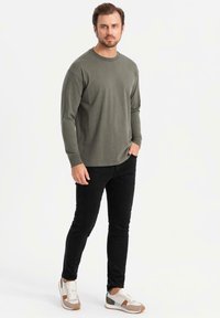 Camicia verde oliva a maniche lunghe, pantaloni neri slim-fit e sneakers multicolori. La camicia ha un collo a giro e polsini a coste.