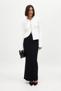 Bela rebrasta majica z dolgimi rokavi s ovratnikom in črnimi gumbi, v kombinaciji z oprijetim črnim maksi krilom; drži črno torbico; čevlji s peto.