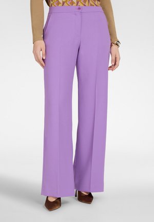 Donna che indossa pantaloni larghi lavanda a vita alta con piega frontale, abbinati a tacchi a punta marroni e top con motivo.