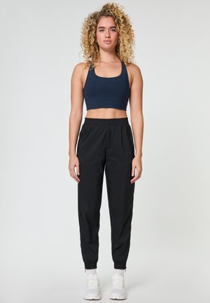 Młoda kobieta o kręconych blond włosach w granatowym sportowym biustonoszu, czarnych spodniach dresowych i białych sneakersach, stojąca na jednolitym tle.