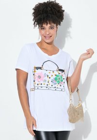 Witte T-shirt met een afbeelding van een handtas met bloemmotieven en gouden studs. Model houdt een kleine metalen handtas vast.