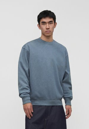 Ung mand med kort sort hår iført en løs blå sweatshirt og mørke bukser, stående mod en ensfarvet lys grå baggrund.
