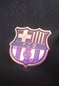 Λαμπερό έμβλημα της FC Barcelona με σταυρό, ρίγες και μπάλα πάνω σε σκοτεινό υφασμάτινο φόντο με υφή, που αντανακλά ροζ και μοβ φως.