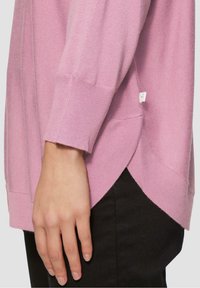 Maglione rosa chiaro con texture a costine, fessure laterali e un'etichetta sull'orlo. Le maniche sono leggermente affusolate e mostrano una mano appoggiata di lato.