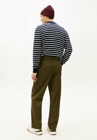 Gestreepte marineblauwe en witte lange mouwen shirt, olijfgroene broeken en witte sneakers met groene accenten. Model is met de rug naar de kijker, draagt een bordeauxrode beanie.