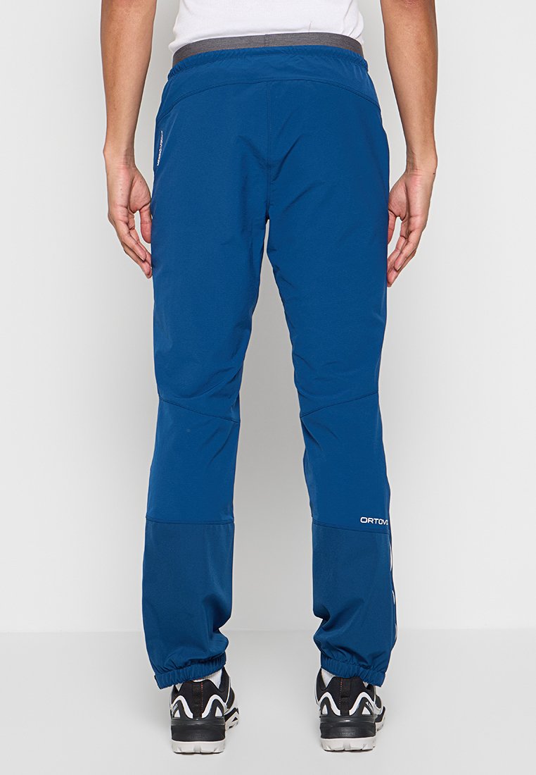 Personne portant un pantalon bleu à taille élastique et des baskets noir et blanc, se tenant dos à la caméra contre un fond uni.