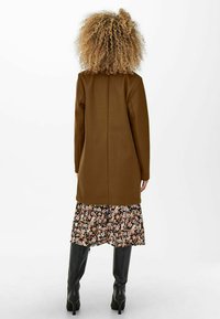 Bruine langwerpige jas met lange mouwen, gedragen over een rok met bloemenpatroon. Gladde textuur, strakke lijnen en kniehoogte zwarte laarzen.