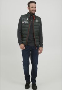 Dunkelgrüne Pufferweste mit gepolsterten Abschnitten, orangen Reißverschlüssen und Logodetails. Getragen über einem grauen Pullover und kombiniert mit dunklen Jeans und Stiefeln.