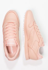 Ett par pastellrosa Reebok Classic-sneakers med vita sulor, sedd uppifrån och från sidan mot en vit bakgrund.