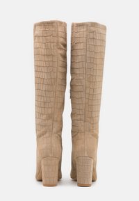Bottes hautes beige en daim texturé avec un motif crocodile, dotées d'un talon épais et d'un design de bout arrondi et lisse.