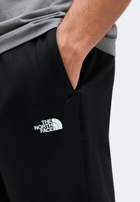 Personne portant un pantalon noir The North Face avec la main dans la poche et un logo visible sur la cuisse, chemise grise partiellement visible.