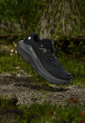 Μαύρο παπούτσι trail running της Salomon με υφή στη σόλα και σύστημα γρήγορου δέσιμου, αιωρούμενο πάνω από διαδρομή με χαλίκια σε δασικό τοπίο.