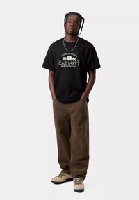 Czarna bawełniana koszulka z nadrukiem, w połączeniu z brązowymi sztruksowymi spodniami i jasnobrązowymi butami. Model stoi z rękami w kieszeniach.