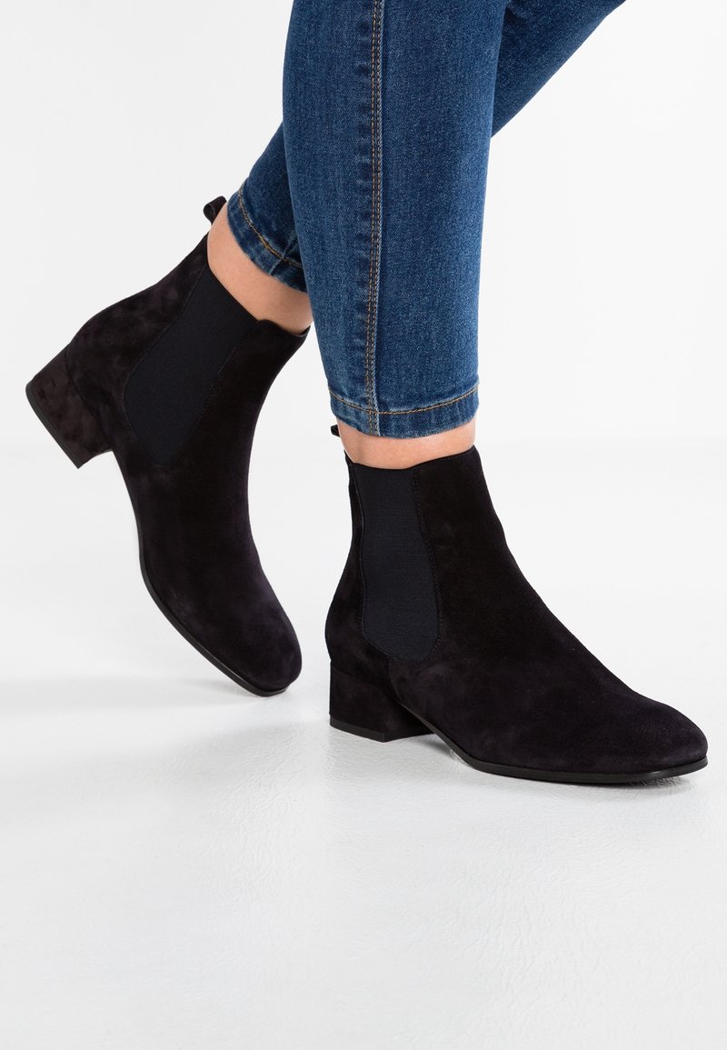 Pieds chaussés de bottines en daim noir avec talons blocs bas et panneaux latéraux élastiques, associées à un jean skinny bleu retroussé aux chevilles.