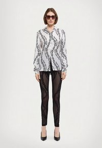 Biała koszula z długim rękawem z czarnym napisem i falistym wzorem, zestawiona z czarnymi legginsami o teksturze koronki oraz czarnymi butami na obcasie ze spiczastym noskiem.