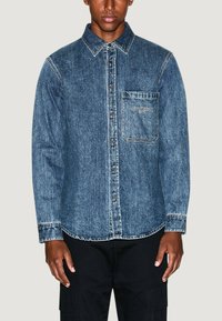 Camicia in denim di colore blu scuro con chiusura a bottoni, colletto a punta e taschino sul petto. Presenta cuciture bianche e un logo sulla tasca.